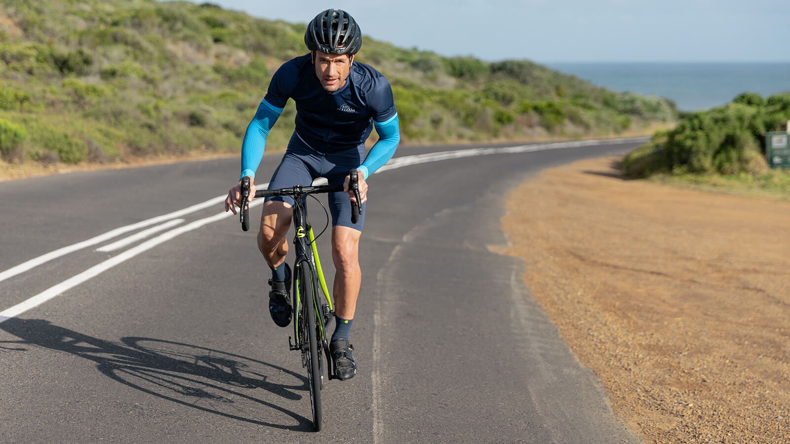 Cyclist wearing Bauerfeind's Sports Compression Arm Sleeves while riding uphill on a paved road surrounded by the ocean and grassy fields.