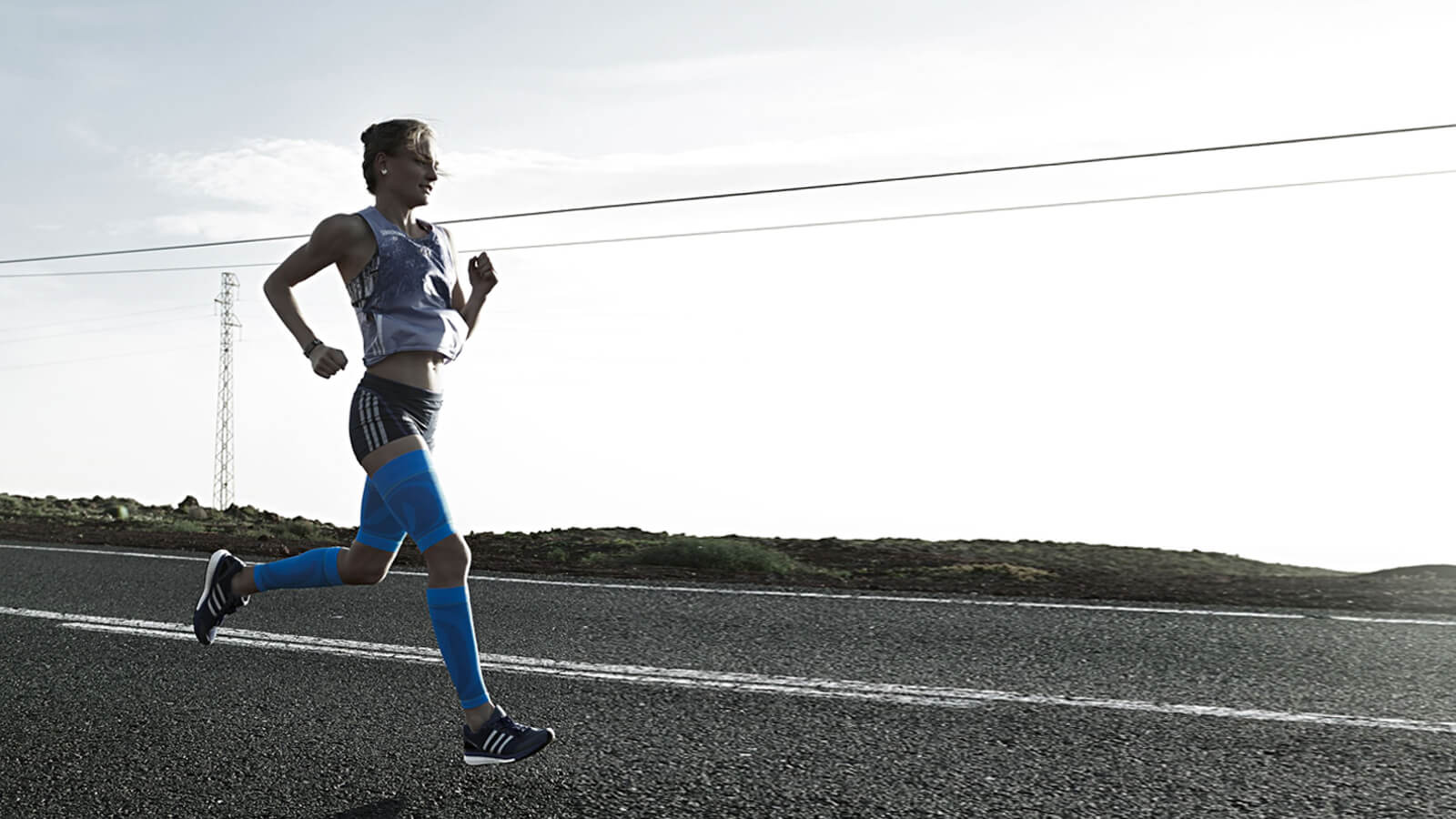 Woman running along a concrete road while wearing Bauerfeind's Sports Compression Upper Leg Sleeves and Sports Compression Lower Leg Sleeves.